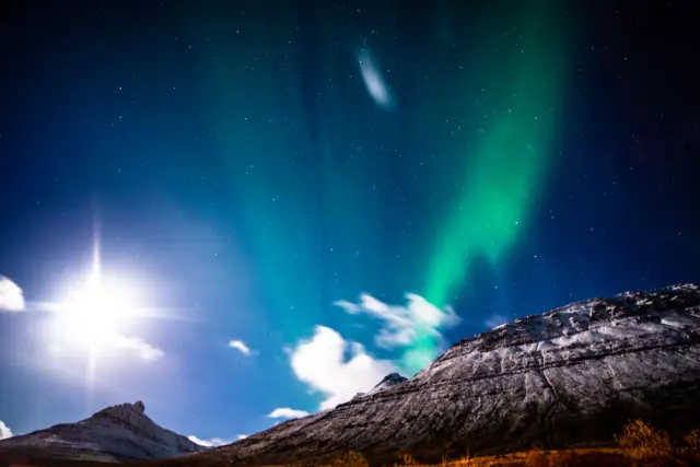 The northern lights streaking across a star-filled sky beside a bright moon over snowy peaks.