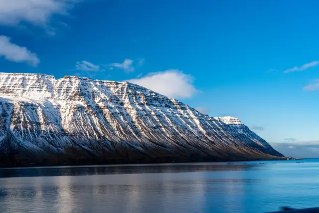 A long snow-streaked mountain ridge catching low sun above a still blue fjord, Iceland