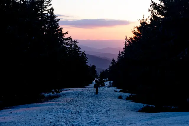 A lone hiker silhouetted on a snowy forest trail overlooking layered mountains at sunset.