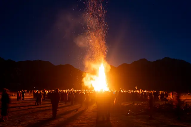A towering bonfire surrounded by a crowd under a starry sky.